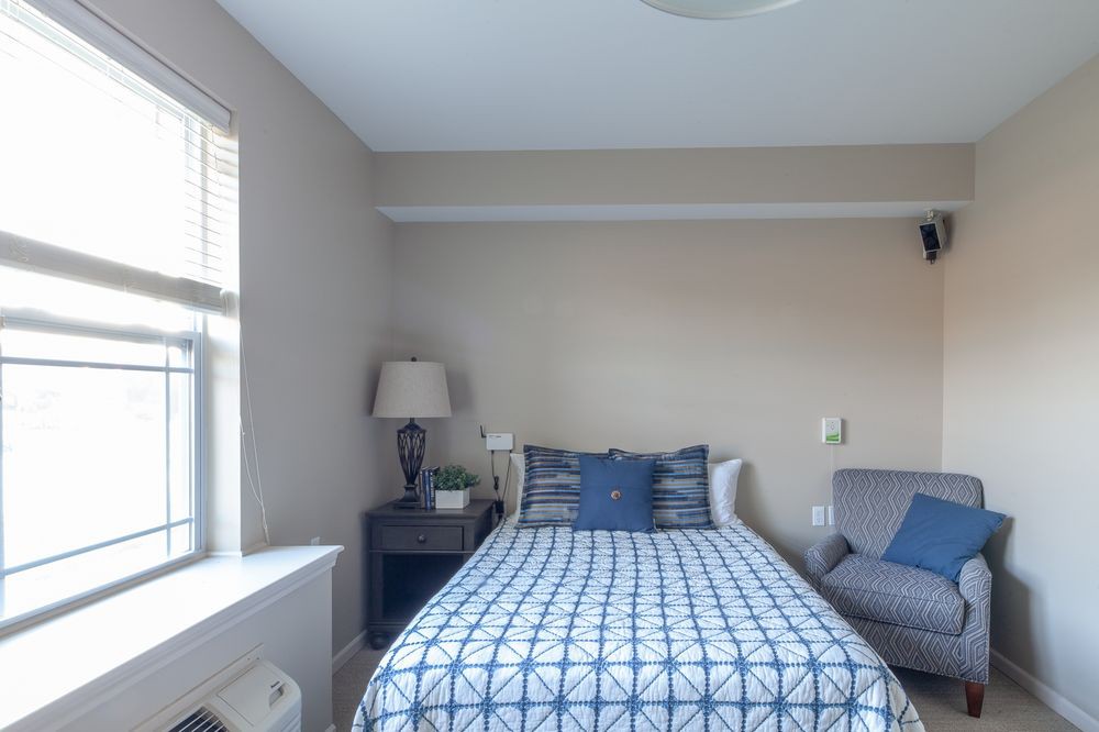 A cozy bedroom with a neatly made bed, a decorative lamp on the nightstand, and a window providing natural light.
