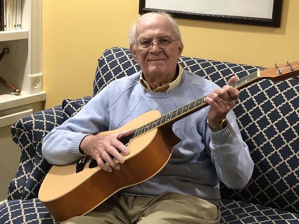 An elderly man sitting on a couch playing an acoustic guitar indoors.