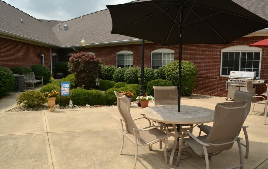 A cozy patio with a table and chairs under an umbrella, surrounded by well-maintained gardens and a brick building in the background.
