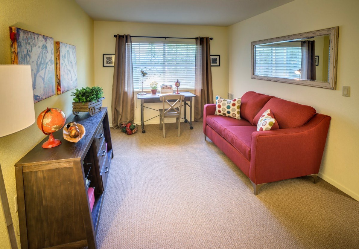 A cozy living room with a red couch, a desk by the window, and decorative shelves.
