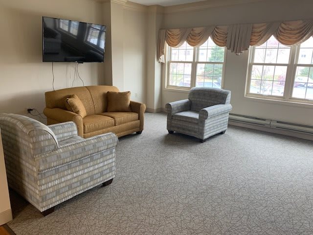 A cozy living room with sofas, an armchair, a wall-mounted TV, and large windows.