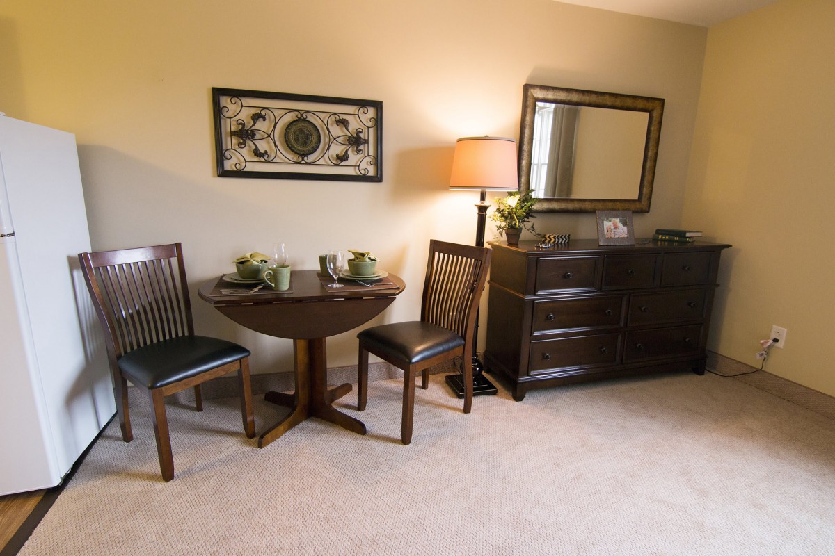 A cozy living room with a small table and two chairs, complemented by a dresser, mirror, and decorative items.