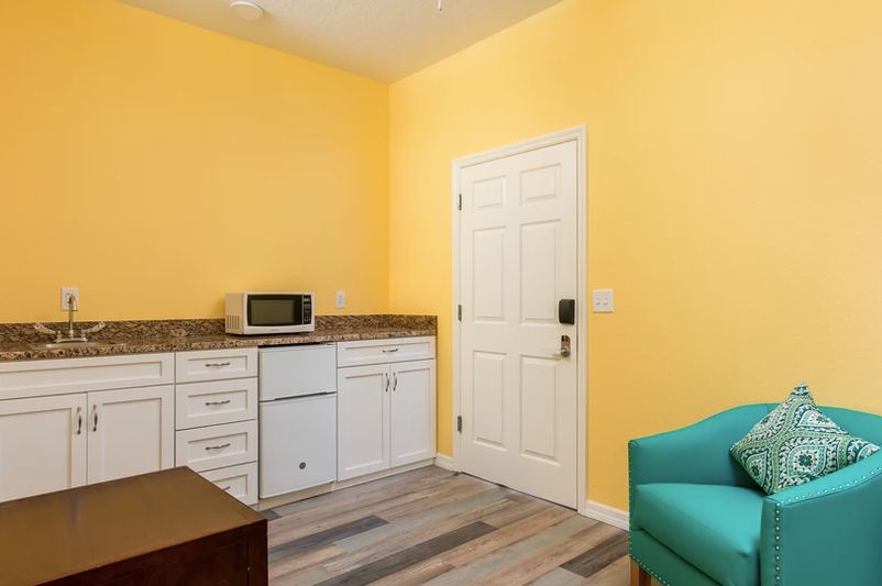A kitchenette area with yellow walls, featuring white cabinets, a microwave, and wood flooring.