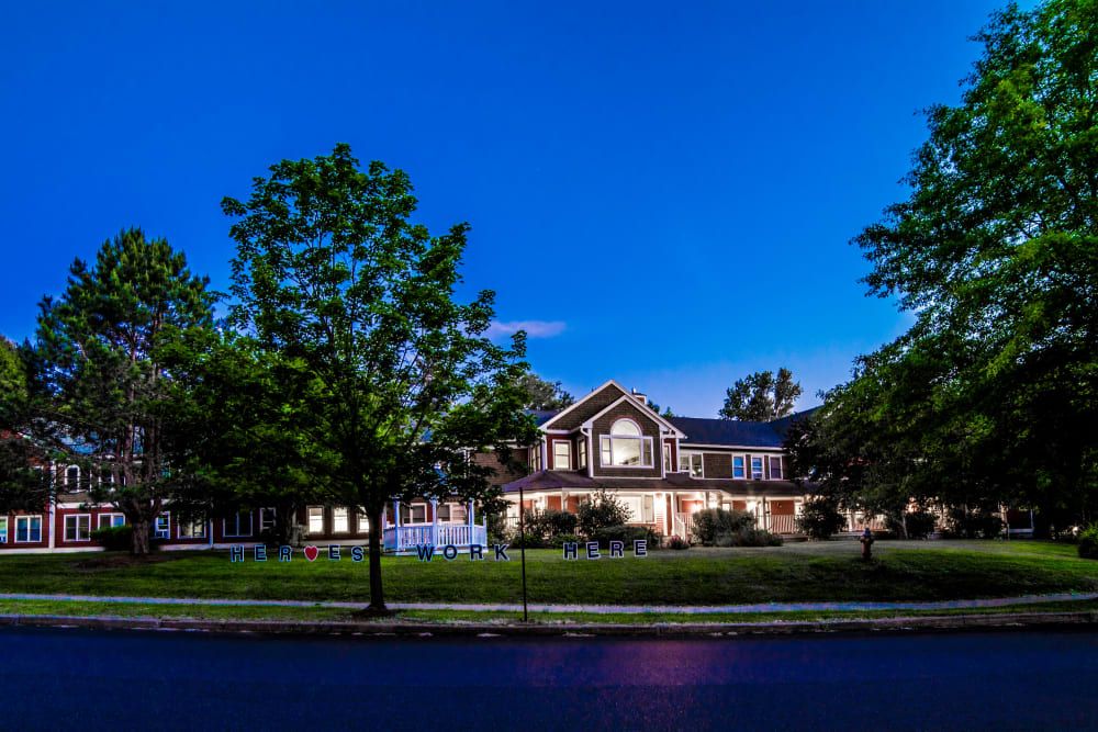 A well-lit residential house in the evening, surrounded by trees and a spacious lawn.