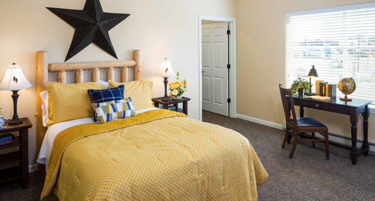A cozy bedroom with a yellow bedspread, decorative pillows, a black star on the wall, and a nightstand with a lamp.