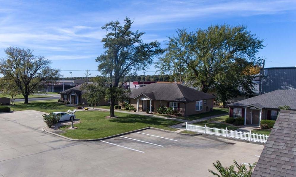 A small building surrounded by a parking lot, trees, and grass under a blue sky with clouds.