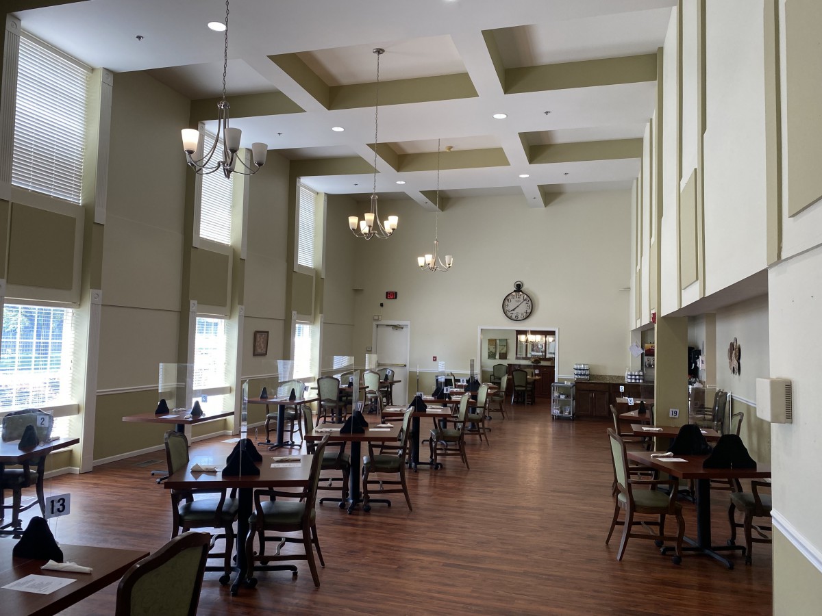 A spacious dining hall with high ceilings, featuring multiple tables and chairs, large windows, chandeliers, and a wooden floor.