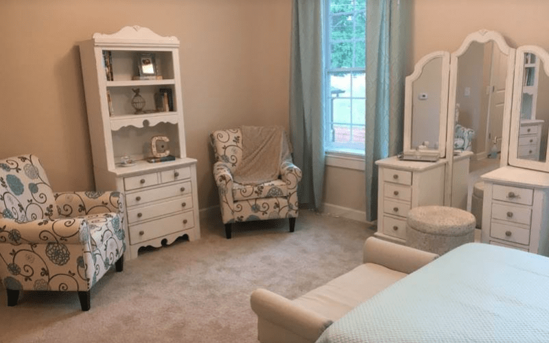 A cozy nursery room with vintage-style furniture and pastel colors, featuring an armchair, dresser, and vanity mirror.