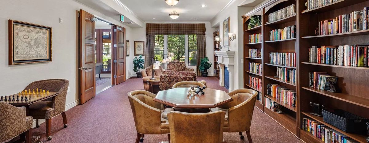 A cozy living room with a table and chairs, surrounded by bookshelves and large windows.