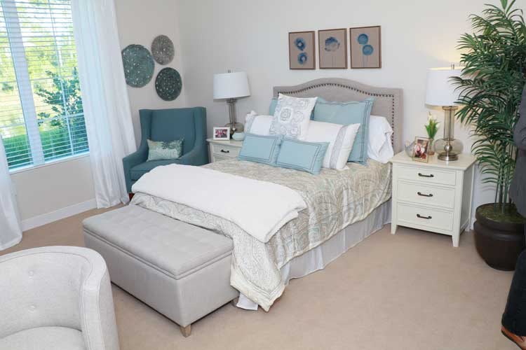 A cozy and elegantly decorated bedroom featuring a neatly made bed with pastel-colored pillows, flanked by bedside tables with lamps, and complemented by wall art and an upholstered ottoman.