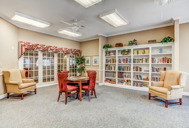 A cozy library room with bookshelves, a table surrounded by chairs, and decorative indoor plants.