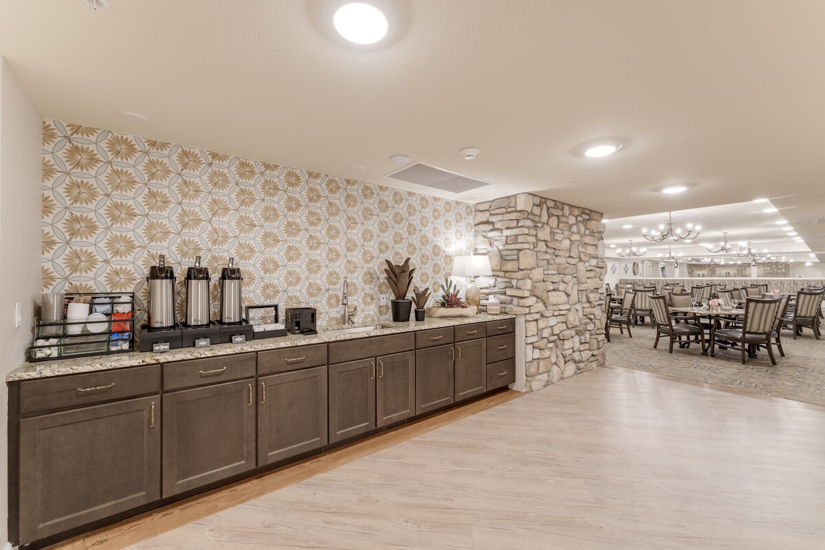 A modern kitchen with a countertop featuring coffee dispensers, leading to a dining area with a stone wall accent.