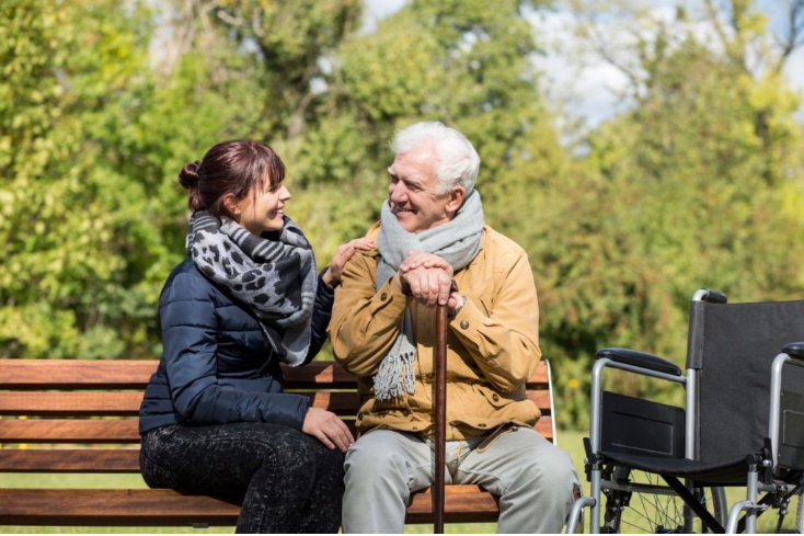 An elderly man with a cane and a younger woman sit on a park bench having a conversation, with a wheelchair nearby.