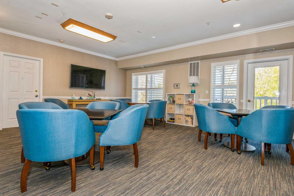 A spacious and well-lit room with blue chairs, several tables, a television, and large windows.