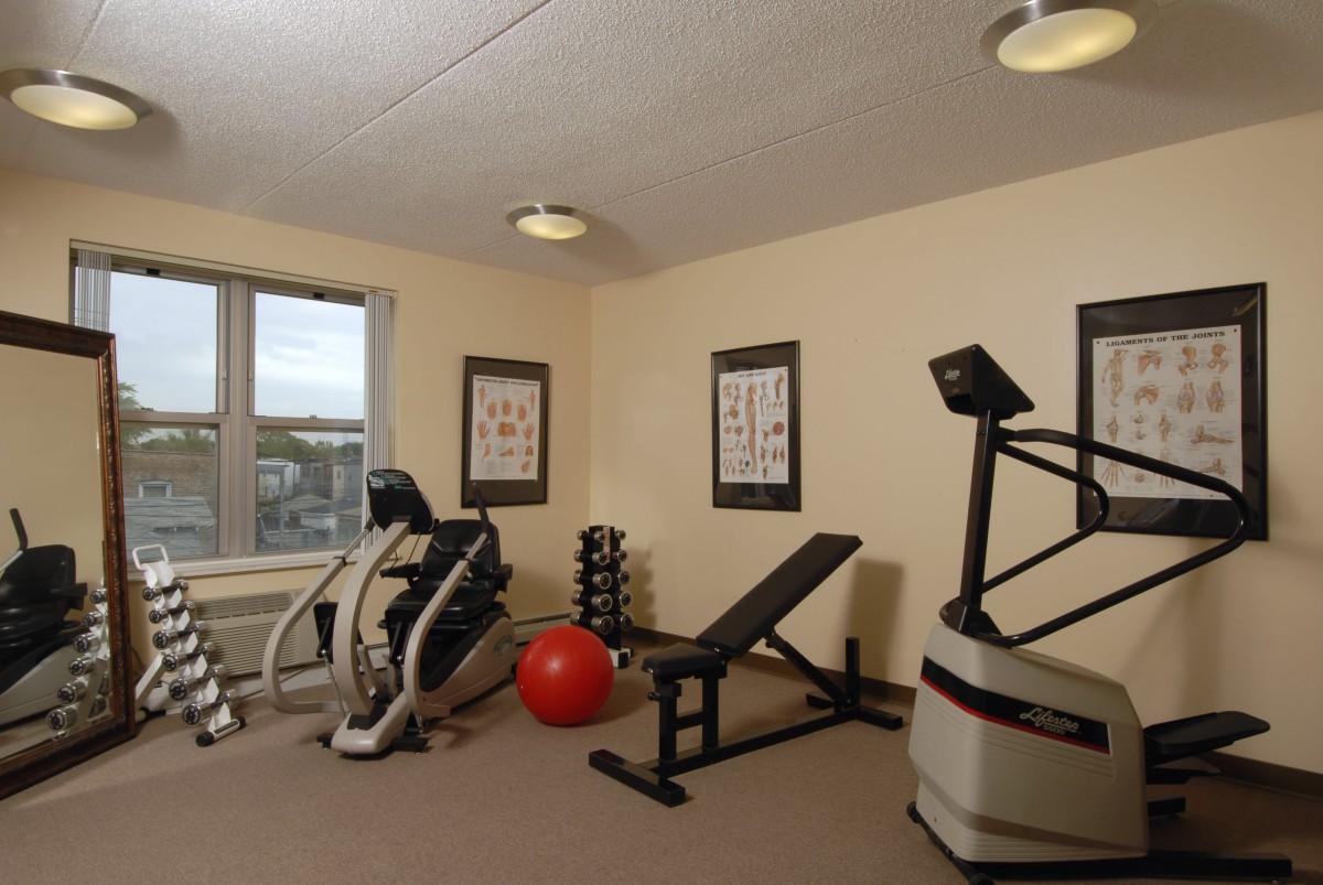 A small gym with various exercise equipment, including an elliptical machine, bench, dumbbells, and a red exercise ball.