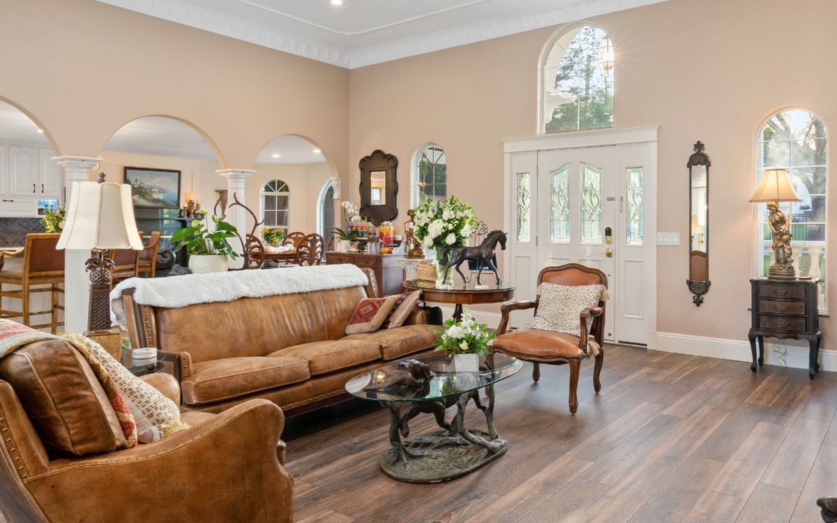 A cozy living room featuring a brown sofa, armchairs, and a glass coffee table adorned with various decorative plants and artwork.