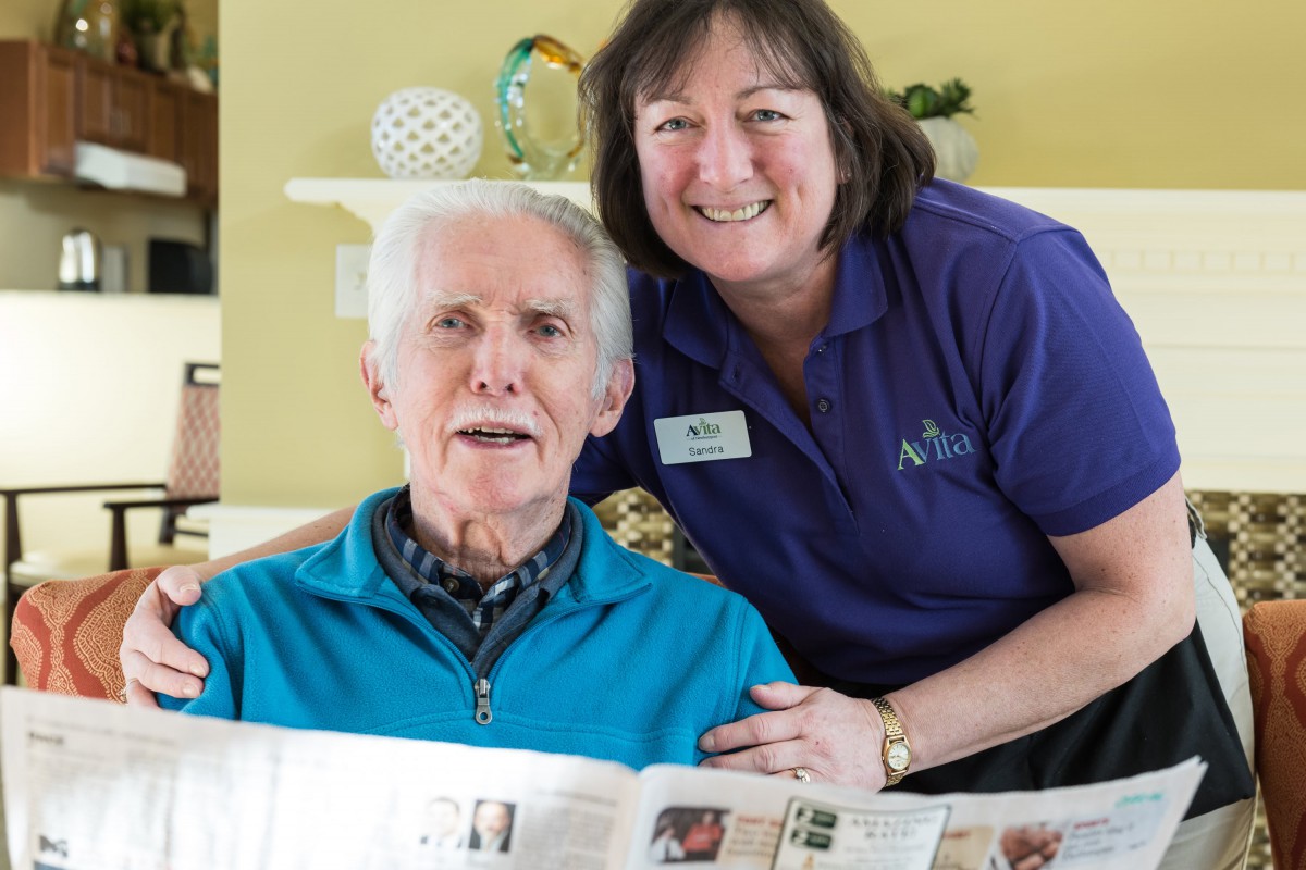 An elderly man and a woman smiling while holding a newspaper indoors.