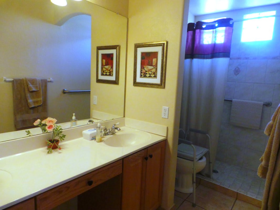 A well-lit bathroom with a large mirror, decorated with artwork and a flower arrangement on the counter.