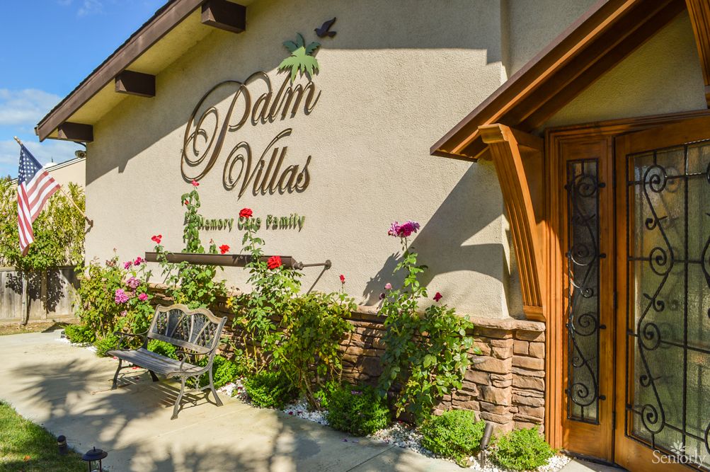 Front view of a building with the sign 'Palm Villas', surrounded by a garden with flowers and plants.
