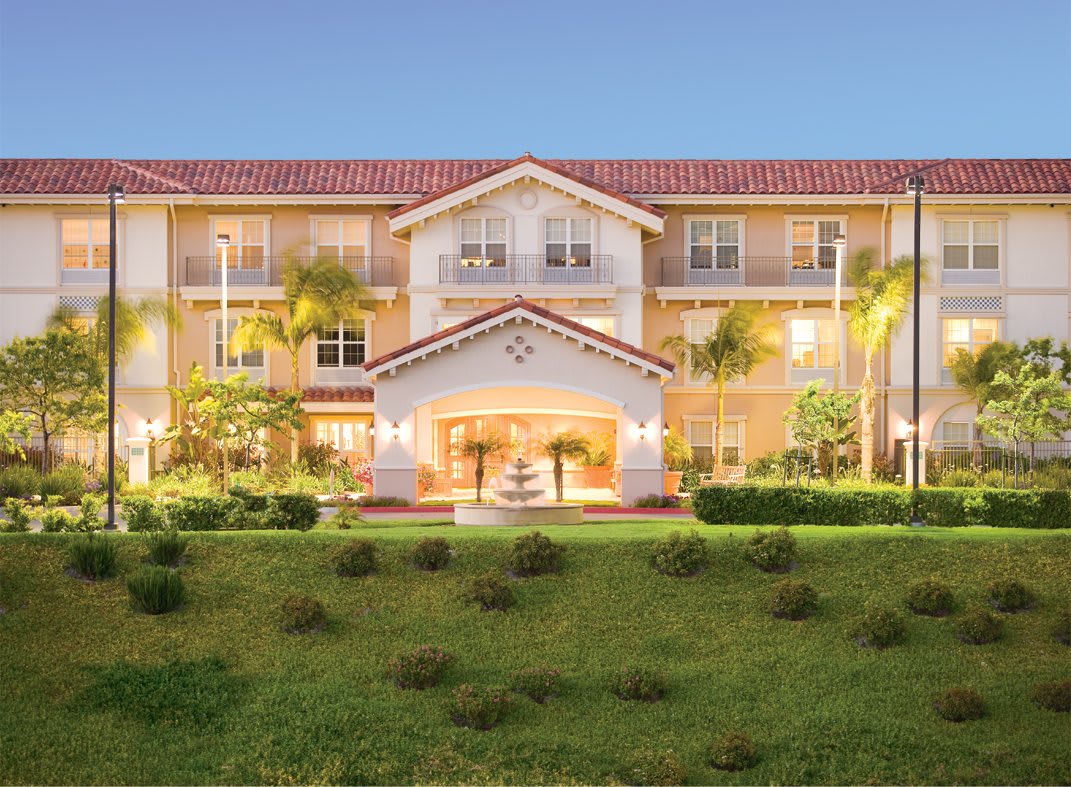 A well-lit residential building with manicured landscaping and palm trees.