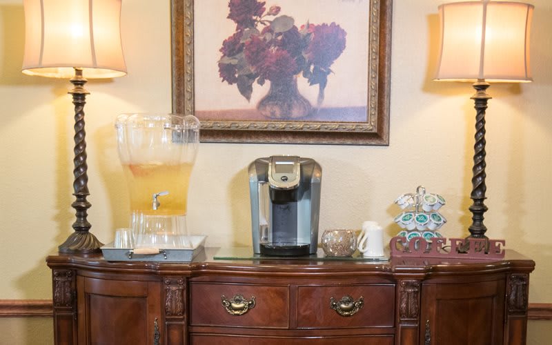 A cozy beverage station featuring a coffee maker and drink dispenser on a wooden sideboard, accented by a framed piece of art.