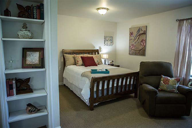 A cozy bedroom with a wooden bed, artwork on the walls, a comfortable armchair, and built-in shelves filled with books.