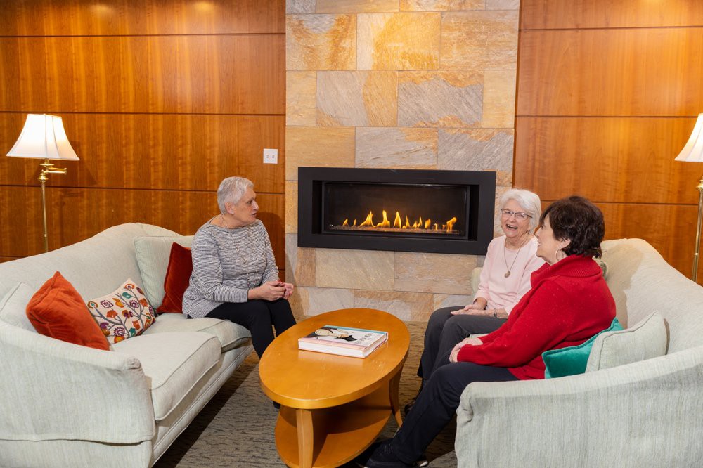 Three seniors enjoy a cozy conversation by the fireplace in a living room setting.