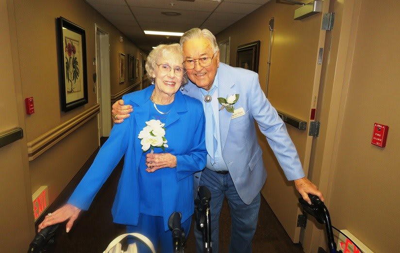 An elderly couple in blue clothing, smiling and posing together in a corridor.