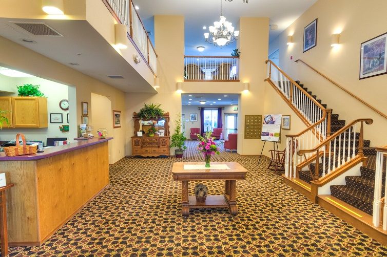 A bright and welcoming hotel lobby featuring a reception desk, elegant chandelier, patterned carpet, and staircase.