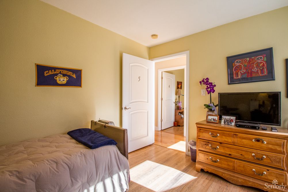 A cozy bedroom featuring a bed, dresser, and floral decorations with warm sunlight streaming in.
