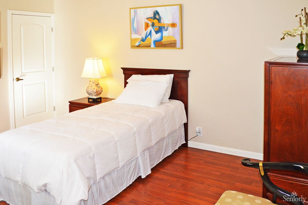A cozy bedroom features a single bed with white linens, a wooden nightstand with a lamp, and artwork on the wall.