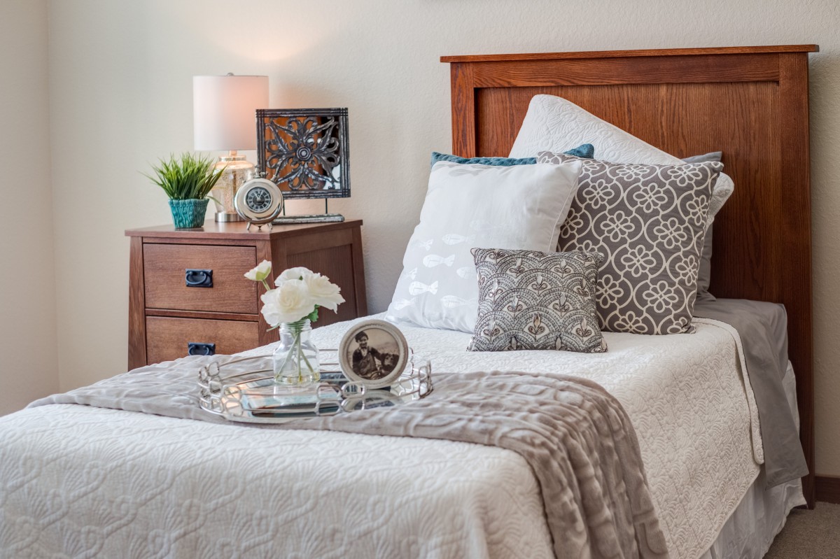 A cozy bedroom with a neatly arranged bed, decorative pillows, and a bedside table featuring a lamp and flowers.