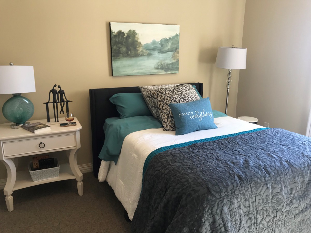 A cozy bedroom with a double bed, featuring decorative pillows and a blue and white color scheme. A painting hangs above the bed, complemented by a nightstand and lamp.