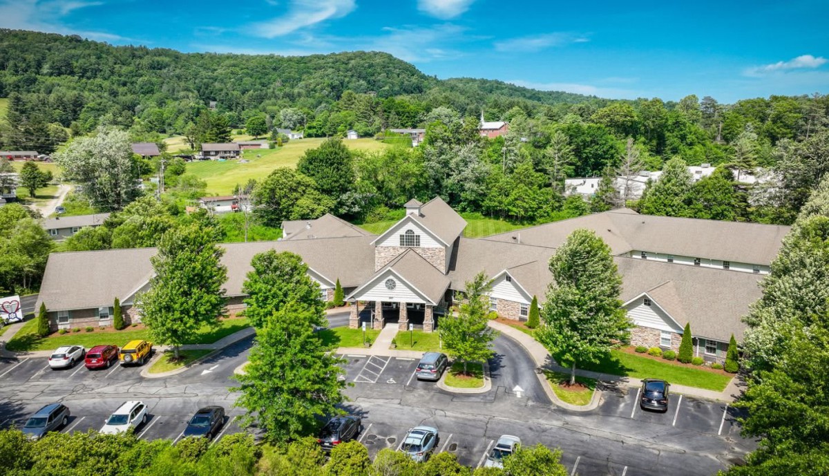 Aerial view of a building surrounded by lush greenery and trees, with a parking lot in the foreground.