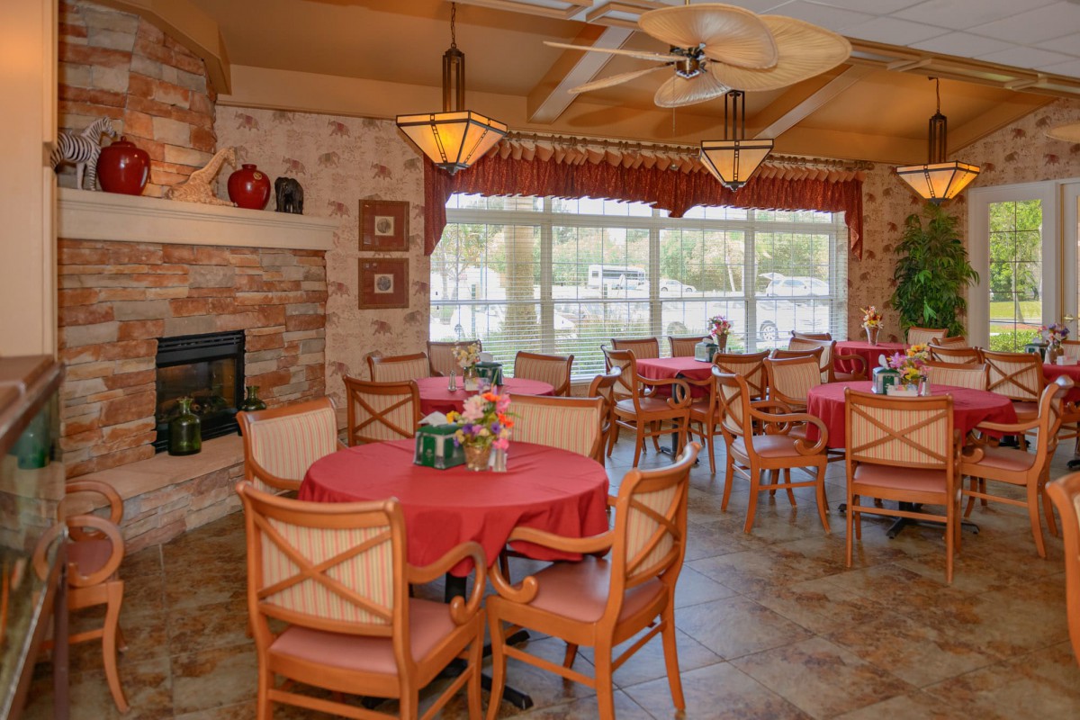 A cozy dining area with multiple tables and chairs, warm lighting, a fireplace, and large windows.