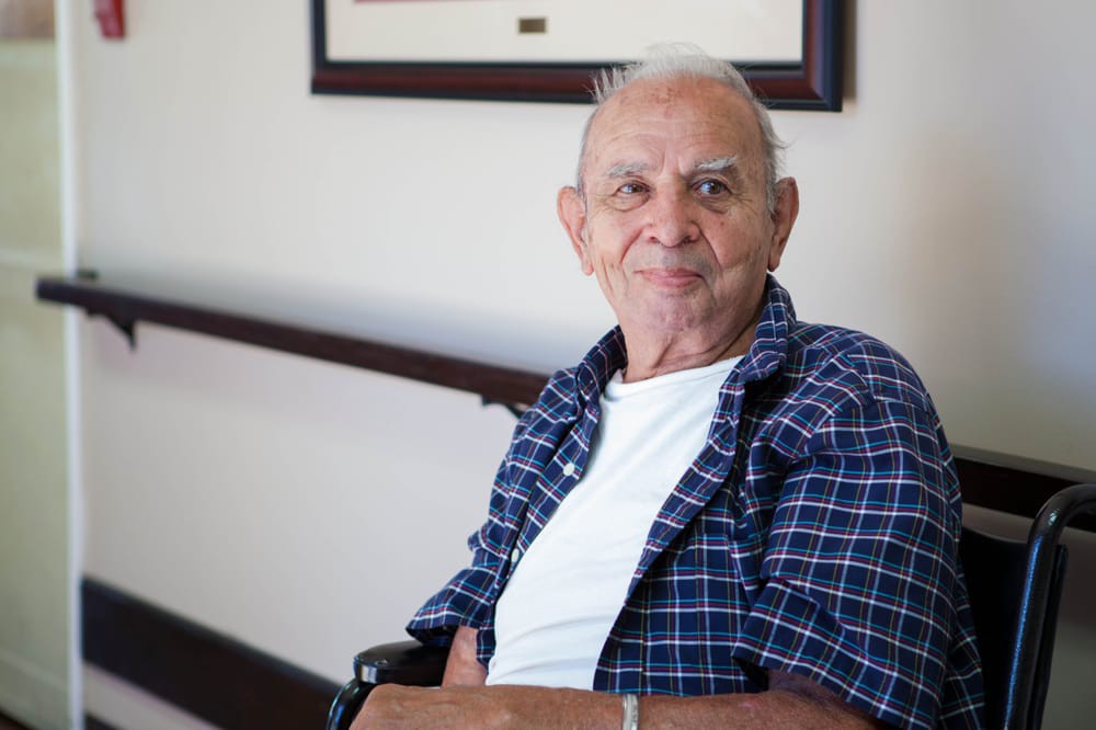 An elderly man sitting in a wheelchair indoors, wearing a blue checkered shirt and a white t-shirt.