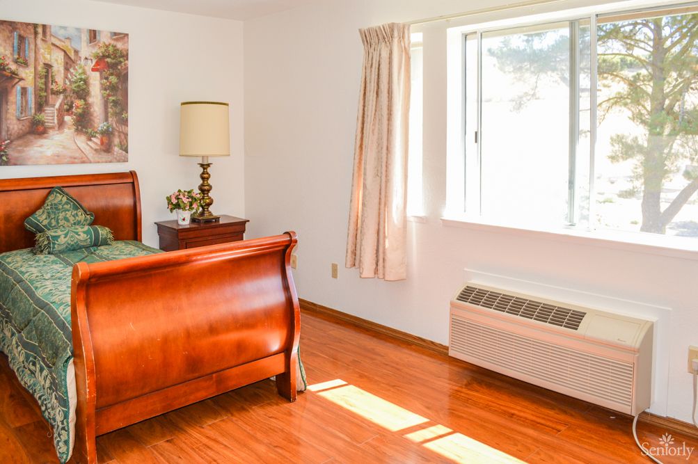 A bright bedroom with hardwood floors, featuring a wooden bed, a lamp on the bedside table, and sunlight streaming through a window with curtains.