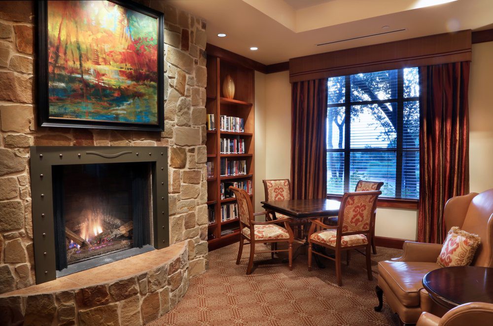 A cozy library nook featuring a stone fireplace, artwork, and a small table with chairs beside a window.