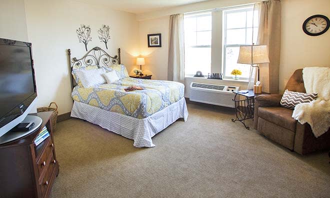 A cozy bedroom with a bed, two lamps, and large windows allowing natural light in.