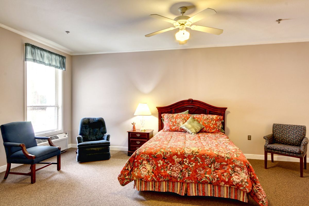 A cozy bedroom featuring a bed with colorful bedding, a bedside lamp, an armchair, and a ceiling fan.