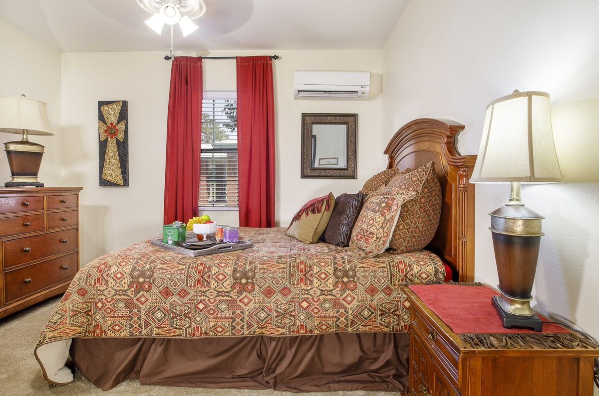 A cozy bedroom featuring a patterned bedspread, wooden furniture, and red curtains, with an air conditioner mounted above the window.