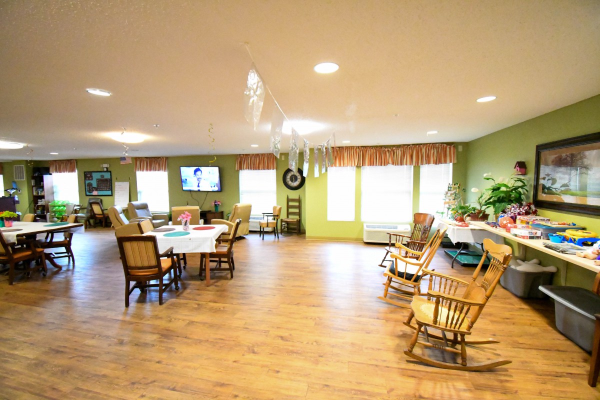 A spacious community room with tables, chairs, and rocking chairs arranged on a wooden floor.