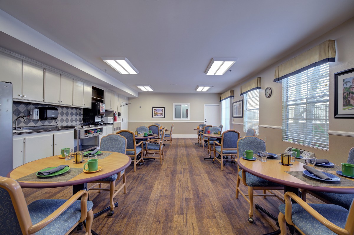 A well-lit dining room with multiple tables and chairs, featuring a wooden floor and a small kitchen area.