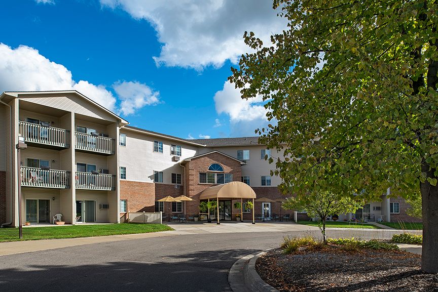 A modern residential apartment building with a driveway and surrounding trees under a clear blue sky.