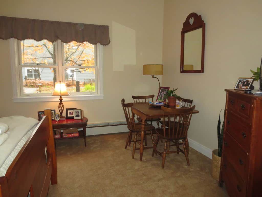 A cozy dining room with a wooden table and chairs, a mirror on the wall, and a lamp by the window.