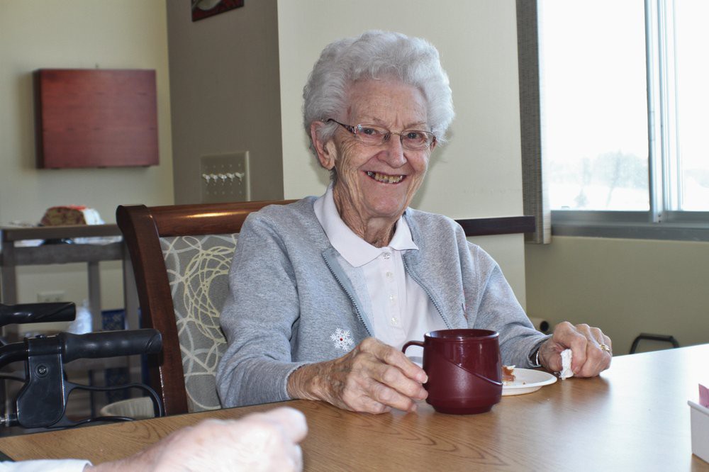 An elderly woman smiling while seated at a table, holding a tea cup.