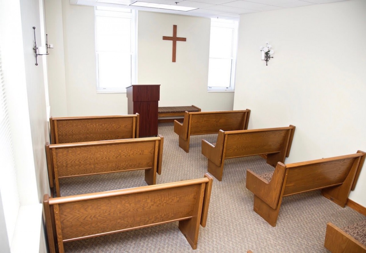A small chapel interior with wooden pews and a pulpit facing a cross on the wall.
