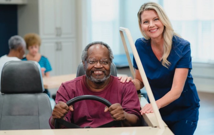 An elderly man smiling while a healthcare worker assists him in a wheelchair simulation.