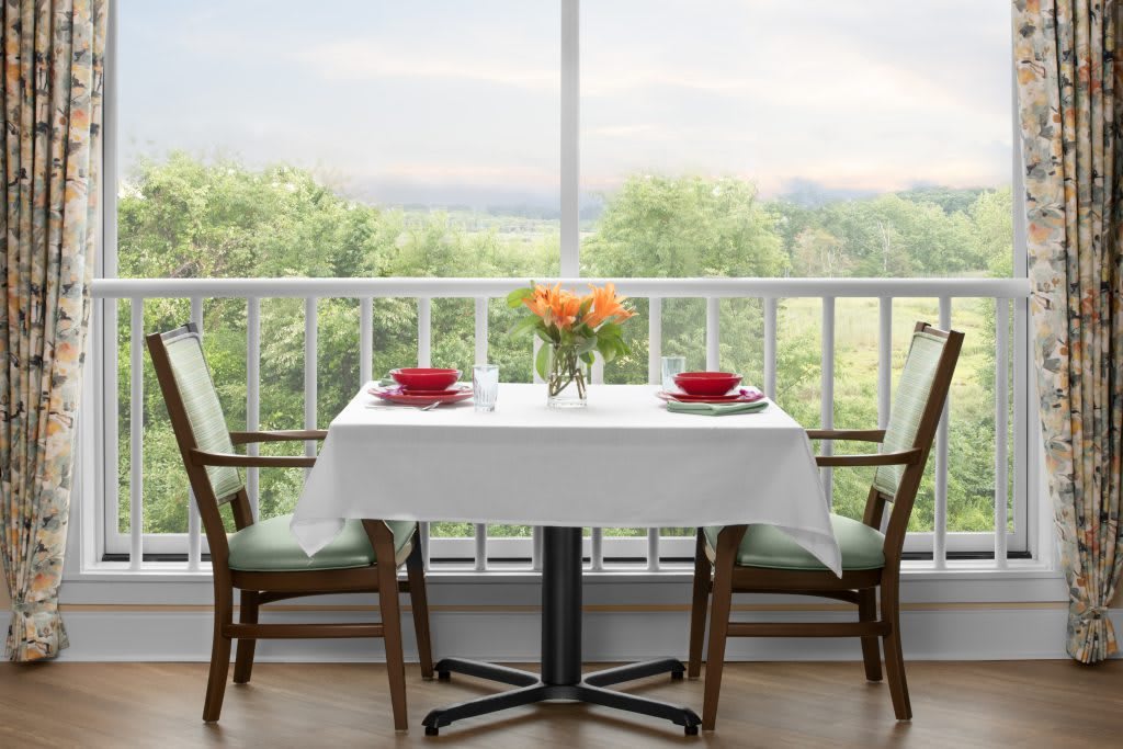 A dining table set for two with red bowls and a vase of flowers by a large window overlooking greenery.