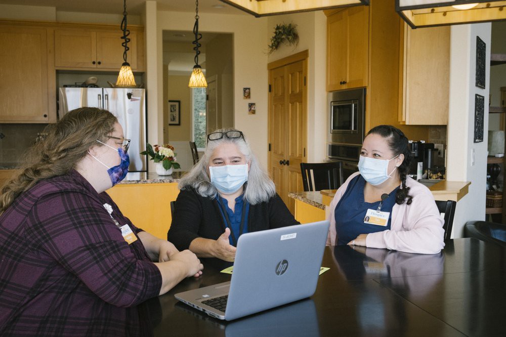 Three people wearing masks are having a discussion around a laptop in a kitchen setting.